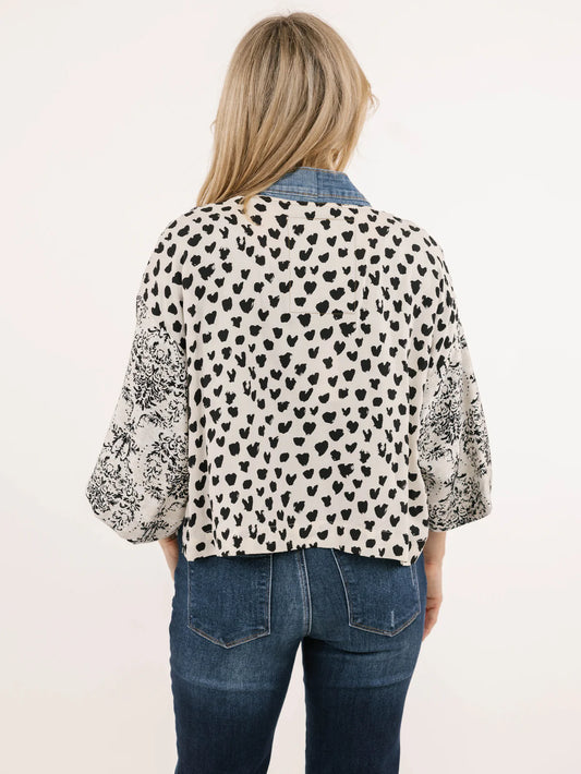 A woman with long blonde hair wears the Shannon Passero Karol Cardigan, a black shirt, and upcycled vintage denim jeans as she stands smiling in front of a textured stone wall, highlighting her sustainable style.