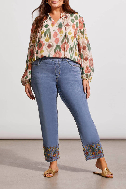 A woman stands wearing a floral print blouse and Tribal's Embroidered Pull On Straight Ankle Jeans with Beading paired with gold sandals.