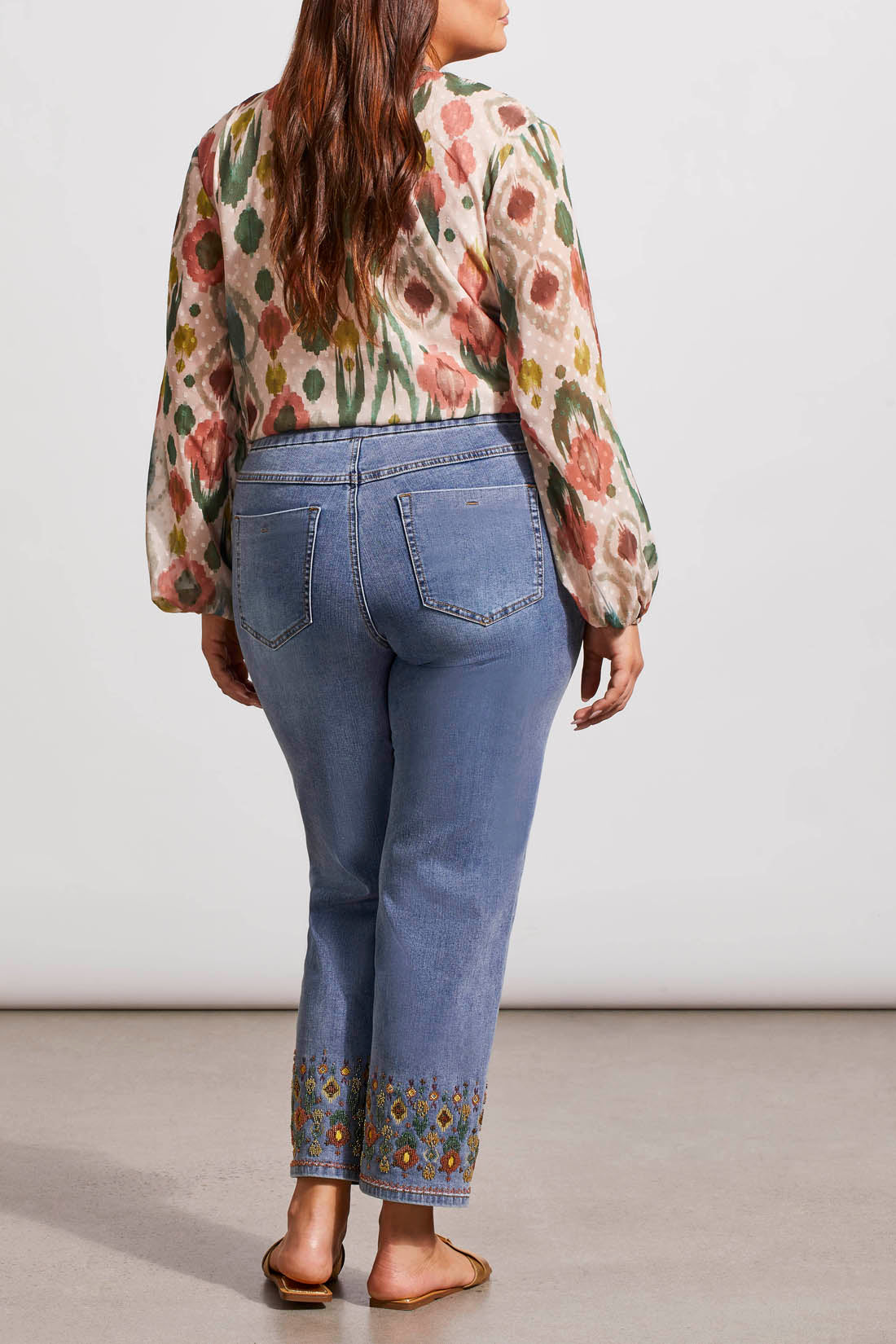 A woman stands wearing a floral print blouse and Tribal's Embroidered Pull On Straight Ankle Jeans with Beading paired with gold sandals.