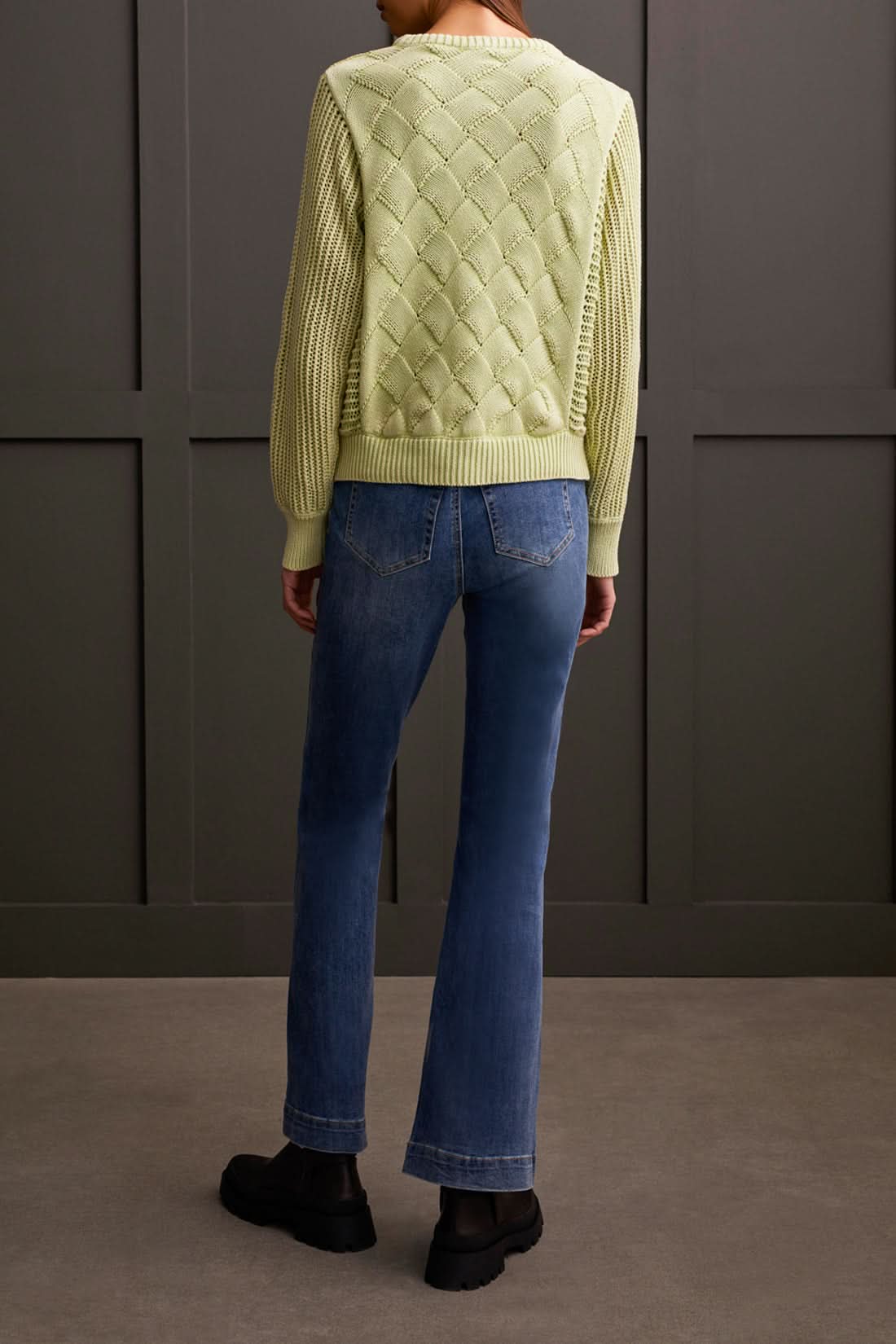 A person is wearing a light green Dolman Crew Neck Sweater with Special Wash from Tribal, along with blue jeans, standing against a dark wall. The luxurious comfort of the soft fabric is evident as the person's hand rests casually in one pocket.