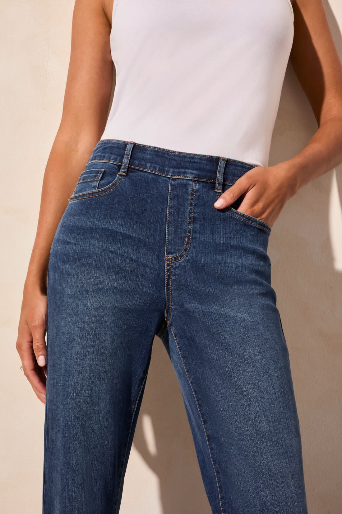 Wearing a white sleeveless top, the person stands against a light background in Tribal's AUDREY PULL-ON FLARE JEANS POCKETS.