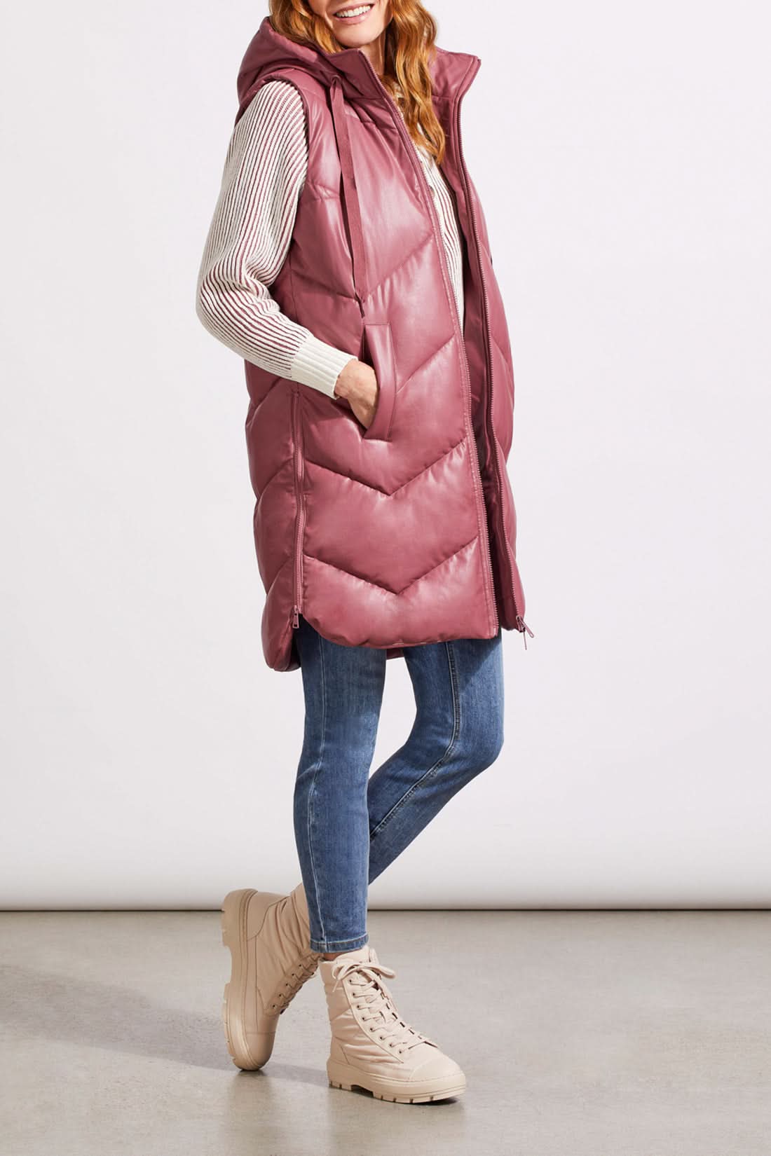A person wearing Tribal's long sleeveless puffer vest in dusty rose over a striped sweater, blue jeans, and beige lace-up boots stands against a plain background.