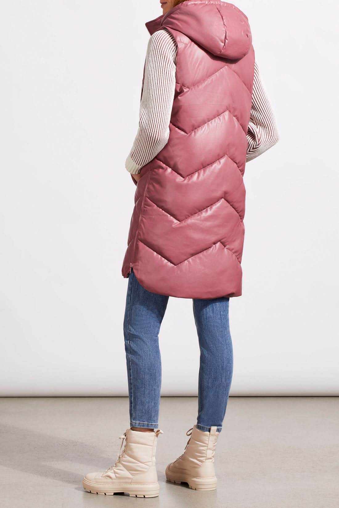 A person wearing Tribal's long sleeveless puffer vest in dusty rose over a striped sweater, blue jeans, and beige lace-up boots stands against a plain background.