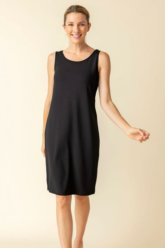 A woman stands smiling in the Habitat Short Everything Dress—a sleeveless black knee-length dress combining comfort and style—ideal for casual occasions, set against a plain light background.