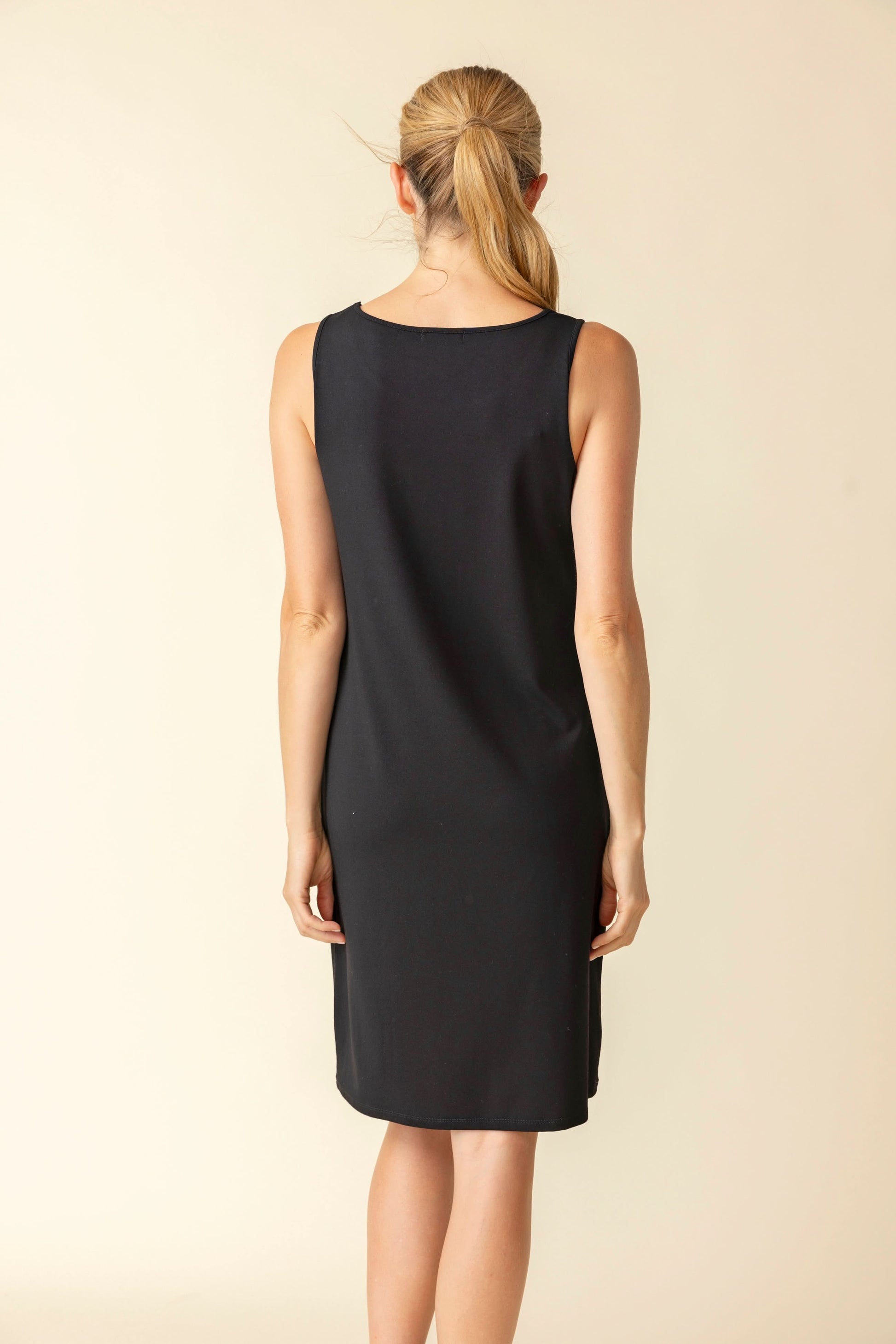 A woman stands smiling in the Habitat Short Everything Dress—a sleeveless black knee-length dress combining comfort and style—ideal for casual occasions, set against a plain light background.