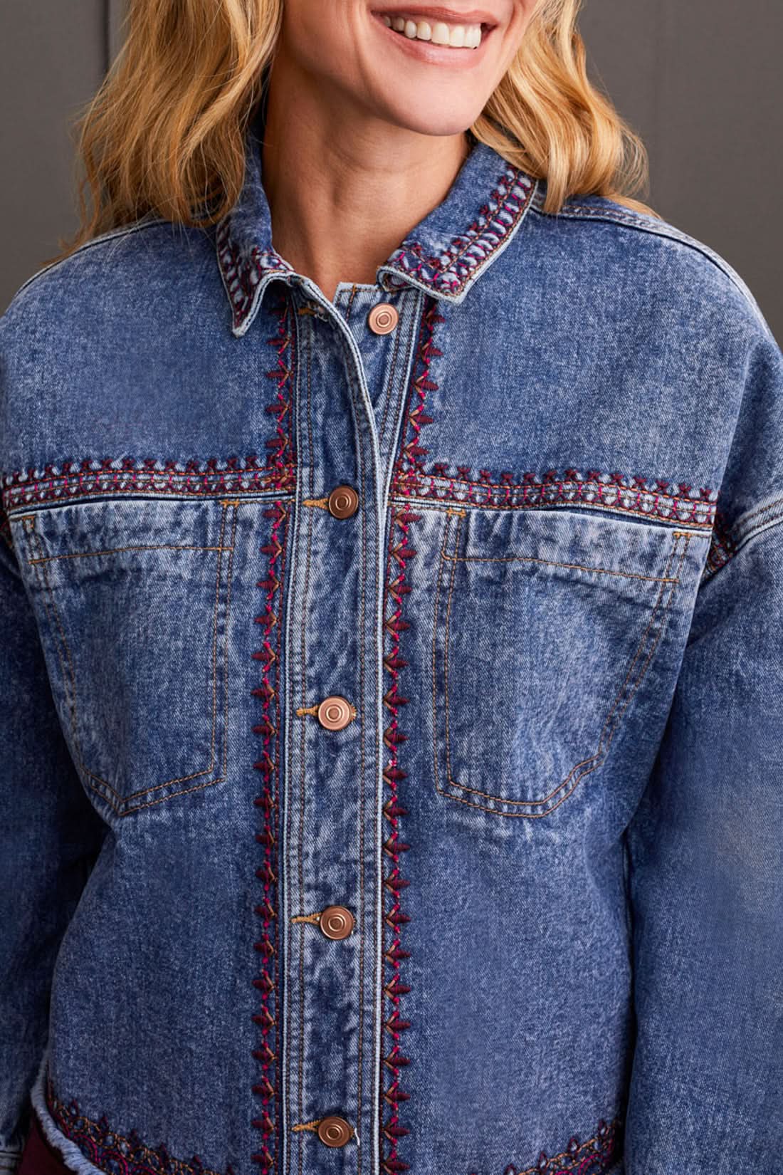 A woman is wearing the Tribal Crop Denim Shacket with Embroidery, featuring a frayed hem, paired with burgundy pants and beige sneakers, standing in front of a dark paneled wall.
