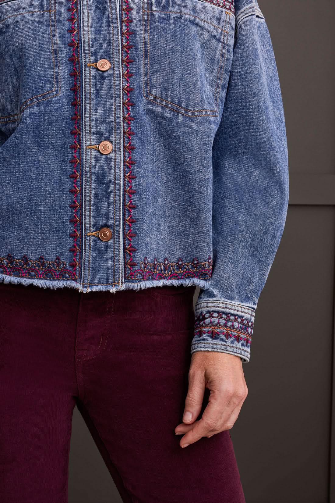 A woman is wearing the Tribal Crop Denim Shacket with Embroidery, featuring a frayed hem, paired with burgundy pants and beige sneakers, standing in front of a dark paneled wall.