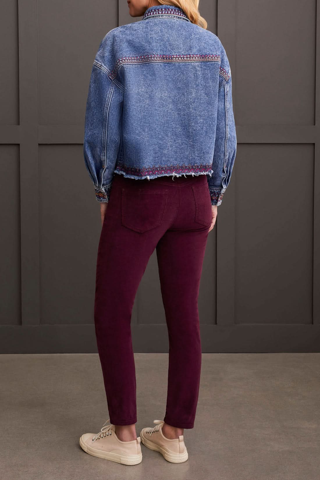 A woman is wearing the Tribal Crop Denim Shacket with Embroidery, featuring a frayed hem, paired with burgundy pants and beige sneakers, standing in front of a dark paneled wall.