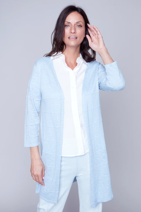 Woman in a light blue Carre Noir knitted cardigan touching her hair, looking to the side against a grey background.