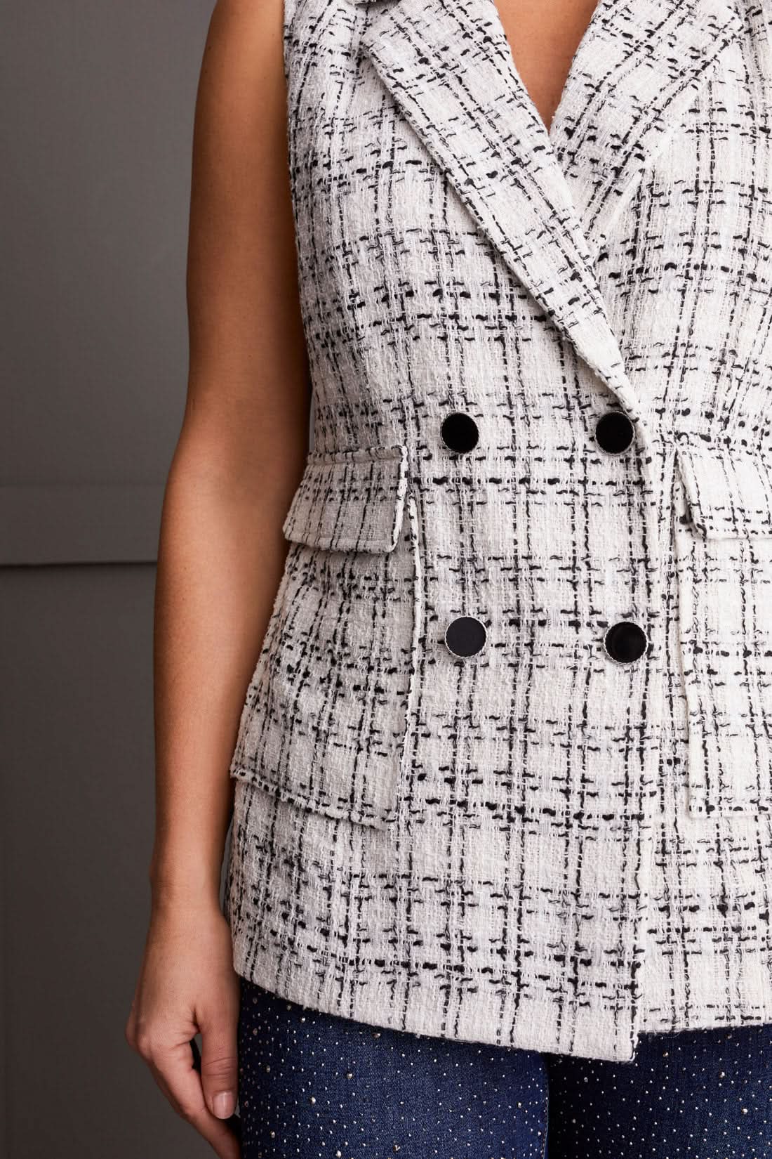 A person wearing the Tribal Lined Double Breast Sleeveless Tweed Vest, dark jeans with a sparkle pattern, and silver ankle boots stands in front of a paneled wall.