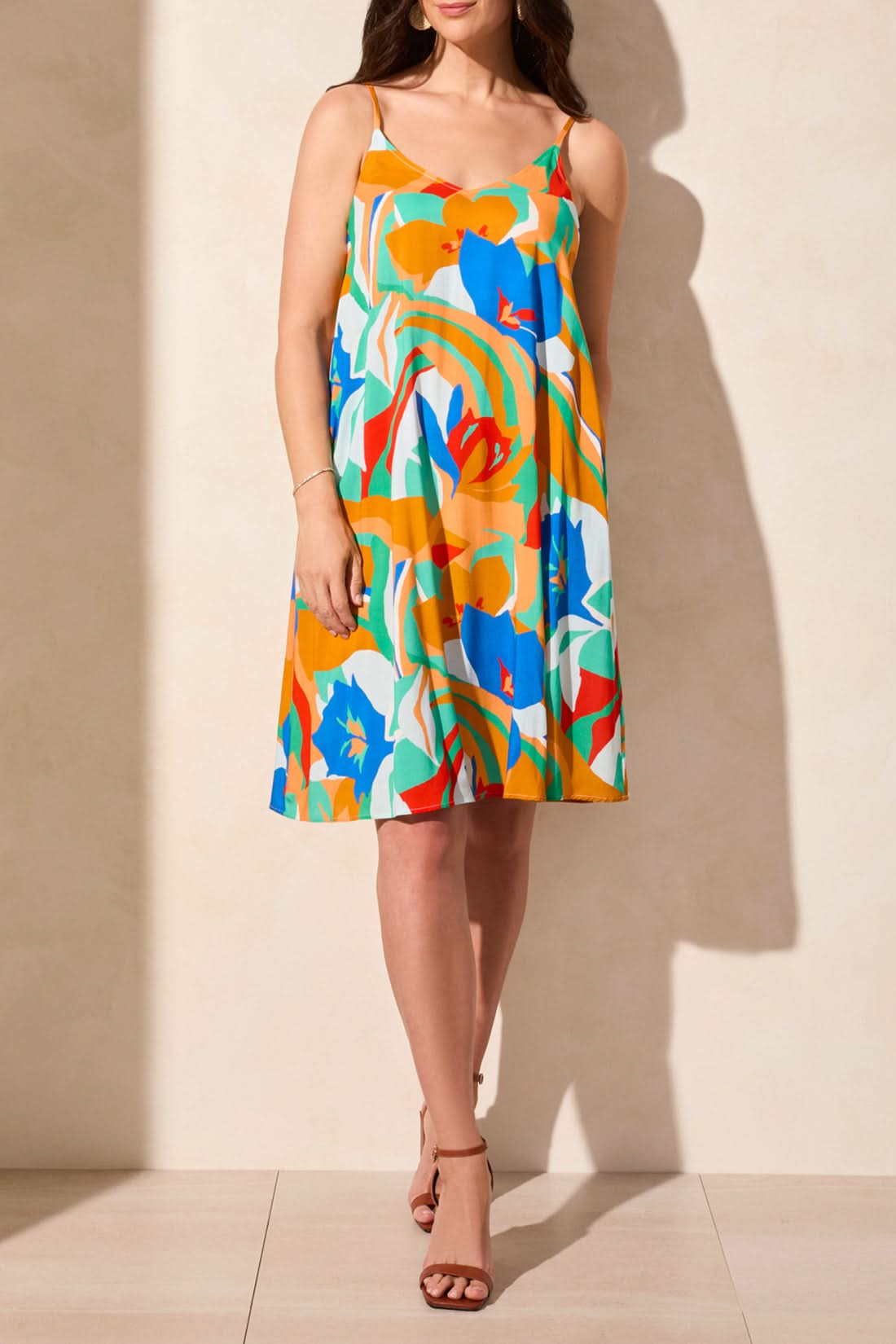 A woman models the Tribal UNLINED DRESS WITH POCKETS, featuring a colorful floral print, thin straps, and sleeveless design, paired with brown ankle-strap sandals against a neutral background.
