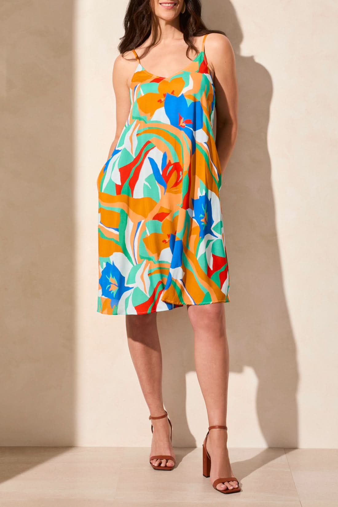 A woman models the Tribal UNLINED DRESS WITH POCKETS, featuring a colorful floral print, thin straps, and sleeveless design, paired with brown ankle-strap sandals against a neutral background.