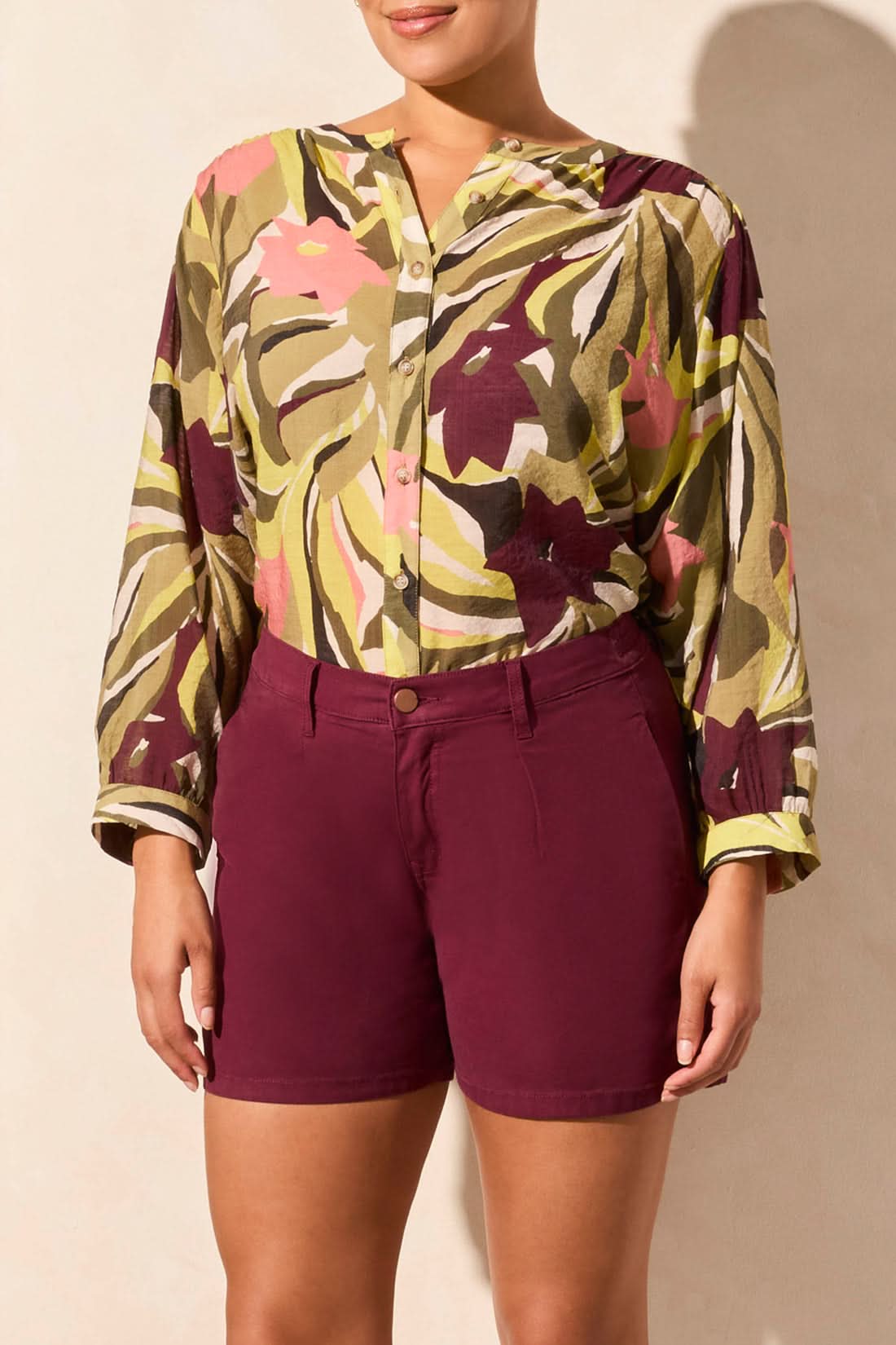 Person poses with one hand in the pocket of Tribal's PLEATED WIDE SHORT, wearing a colorful patterned long sleeve blouse and maroon shorts with handy pockets.