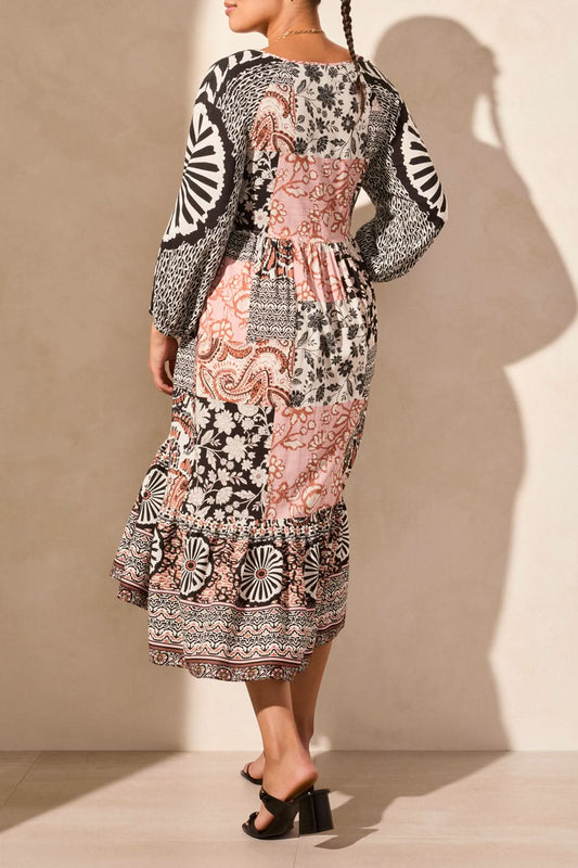 They are elegantly poised on a light floor, wearing the Tribal RAGLAN SLEEVE COMBO PRINT DRESS with 3/4 sleeves and a blend of black, white, and pink floral-geometric designs.