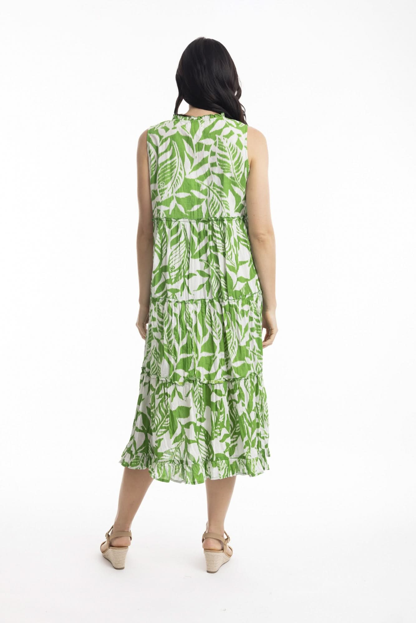 A woman smiles in the Orientique Hayman Green Sleeveless Boho Dress, featuring a green and white leaf pattern and tiered skirt, styled with beige heeled sandals—ideal for summer days against a plain white background.