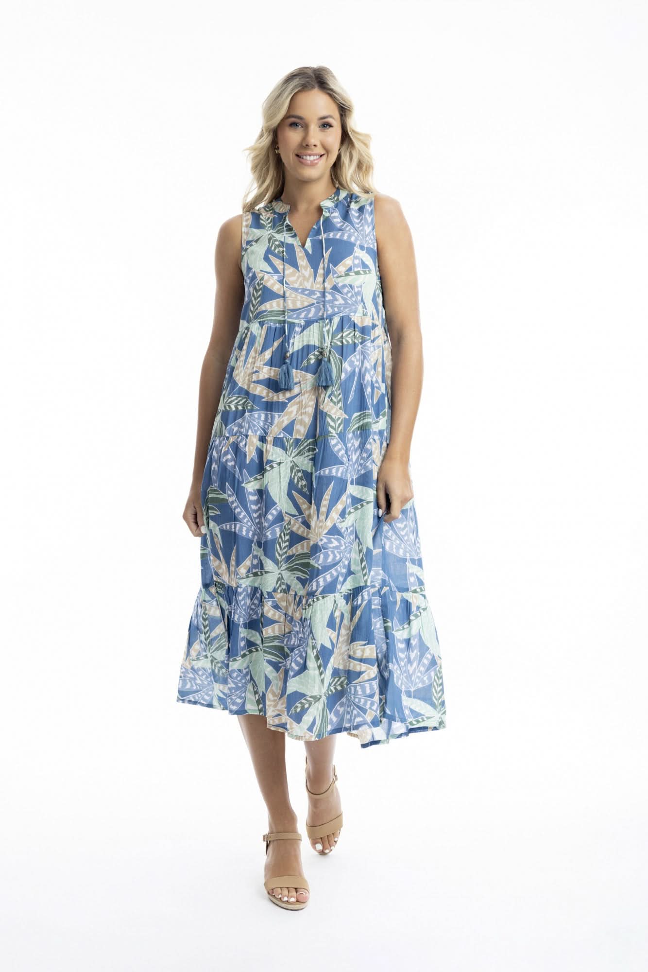 A woman poses against a white backdrop in the Orientique Fitzroy Sleeveless Boho Dress, featuring a blue tropical leaf print and v-neckline, paired with tan wedges—an ideal choice for summer events.
