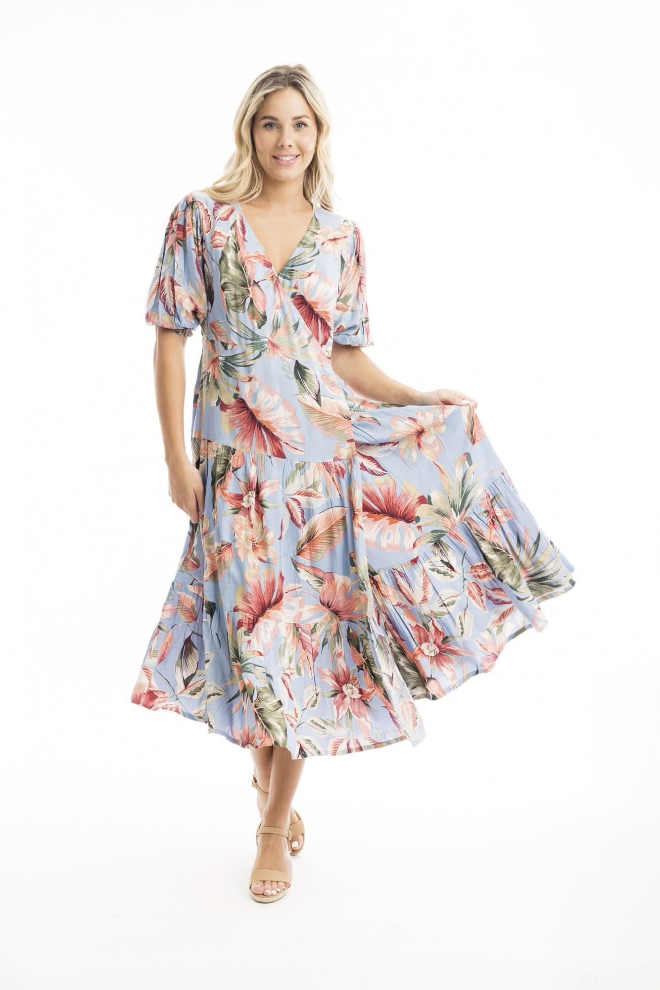 A woman models the Orientique Luzon Midi Crossover Dress—a light blue, floral-patterned summer dress with a v-neckline, short sleeves, and ruffled hem—paired with tan heeled sandals against a white background.