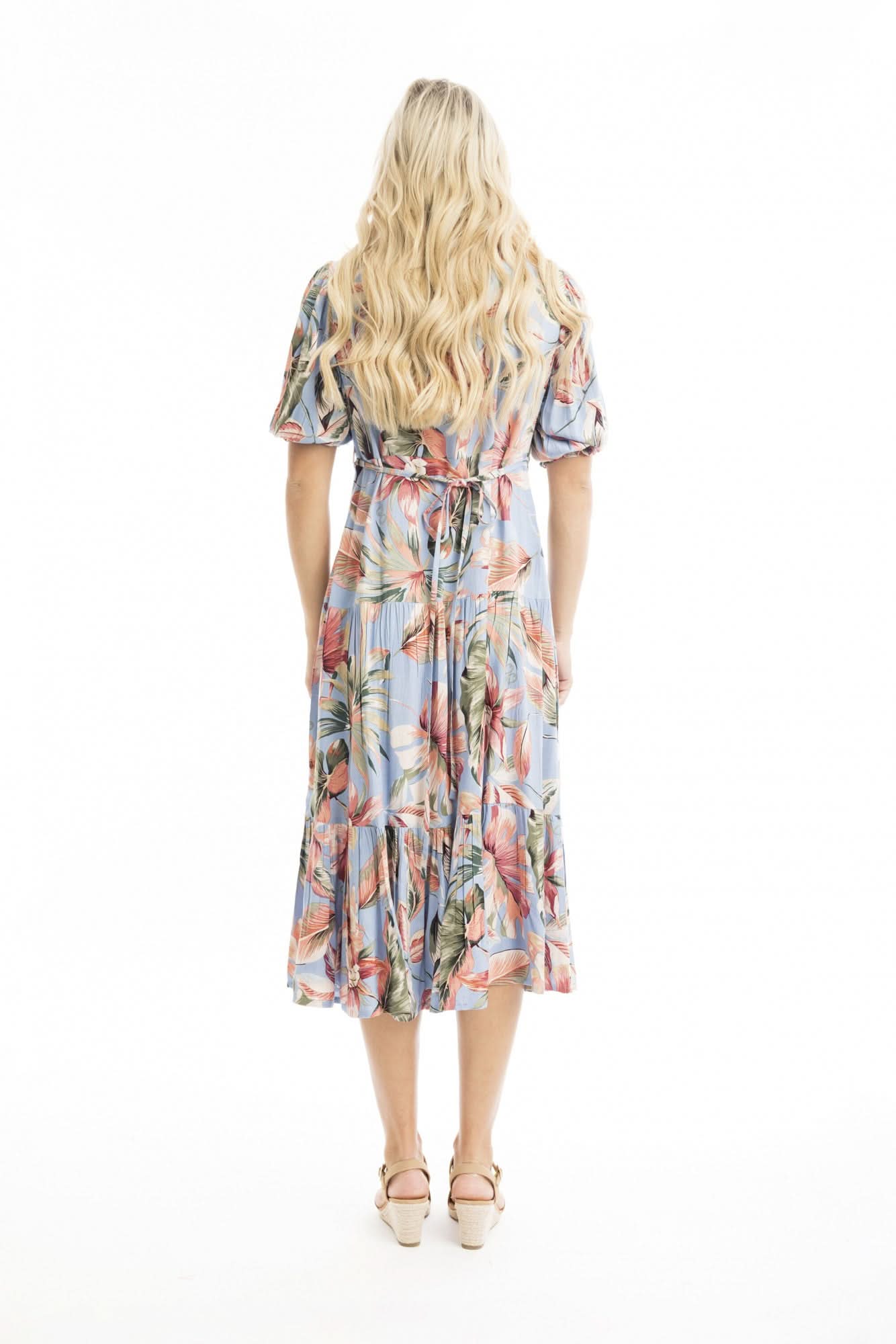 A woman models the Orientique Luzon Midi Crossover Dress—a light blue, floral-patterned summer dress with a v-neckline, short sleeves, and ruffled hem—paired with tan heeled sandals against a white background.