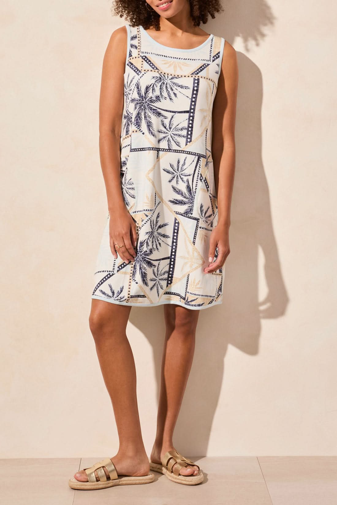 A person wearing Tribal’s REVERSIBLE A-LINE DRESS in a blue and beige leaf pattern stands against a light wall, exuding comfort with a sideways glance and one hand in their pocket.