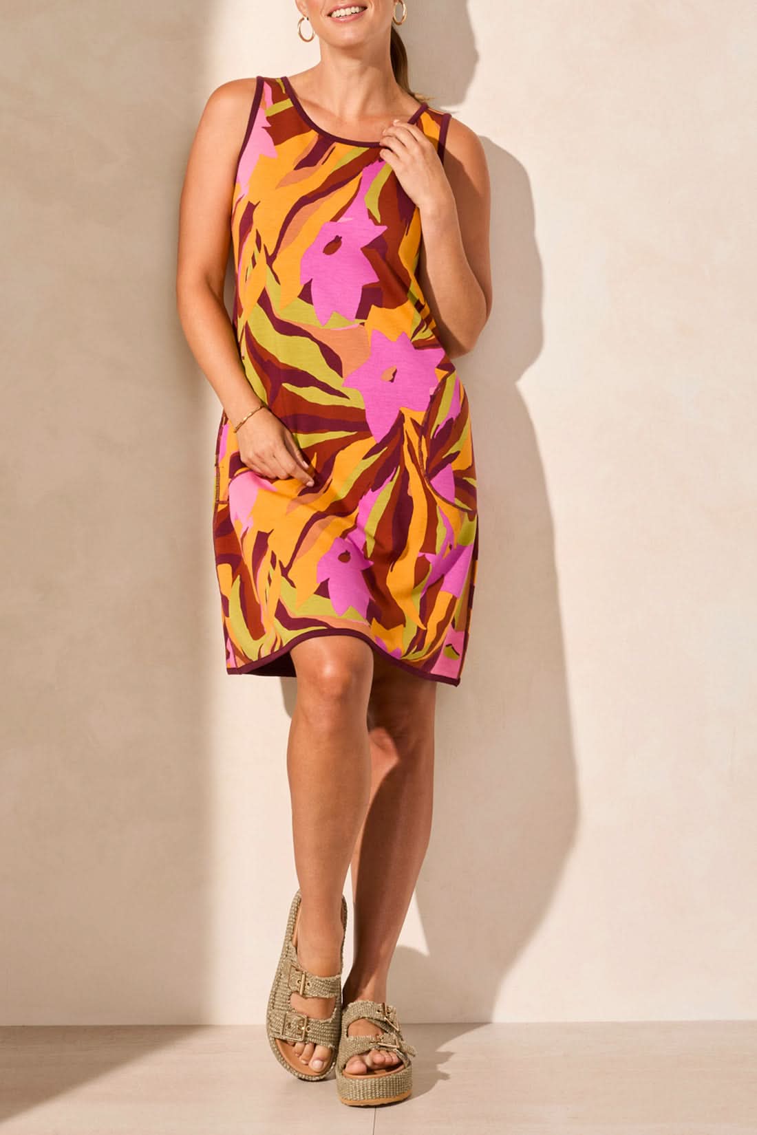 A person wearing Tribal’s REVERSIBLE A-LINE DRESS in a blue and beige leaf pattern stands against a light wall, exuding comfort with a sideways glance and one hand in their pocket.