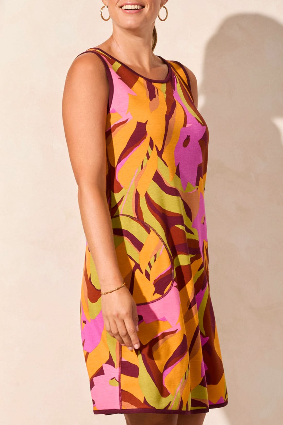 A person wearing Tribal’s REVERSIBLE A-LINE DRESS in a blue and beige leaf pattern stands against a light wall, exuding comfort with a sideways glance and one hand in their pocket.