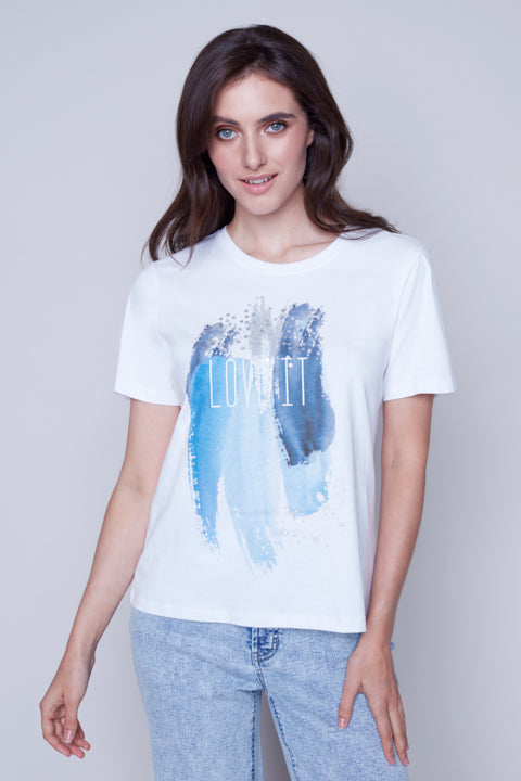 Woman in a Love It Round Neck Tee by Carre Noir featuring text and graphic design, paired with blue jeans, standing against a grey backdrop, perfect for a casual outing.