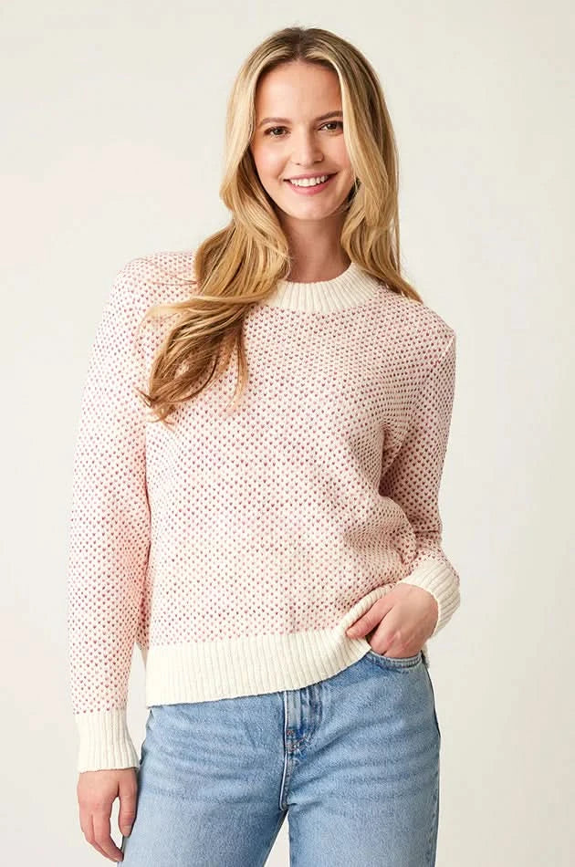 A woman with long hair wears a Parkhurst Kinsley Sweater adorned with heart-shaped polka dots and blue jeans, smiling while facing the camera.