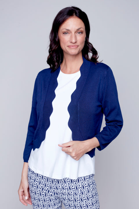 A woman wearing a blue Carre Noir short wavy cardigan, white top, and patterned trousers standing against a gray background.