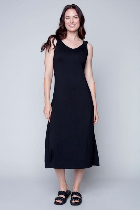 A woman standing against a plain background wearing a Sleeveless Flow Dress by Carre Noir made of soft fabric and embellished sandals.
