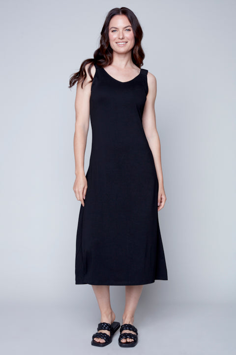 A woman standing against a plain background wearing a Sleeveless Flow Dress by Carre Noir made of soft fabric and embellished sandals.
