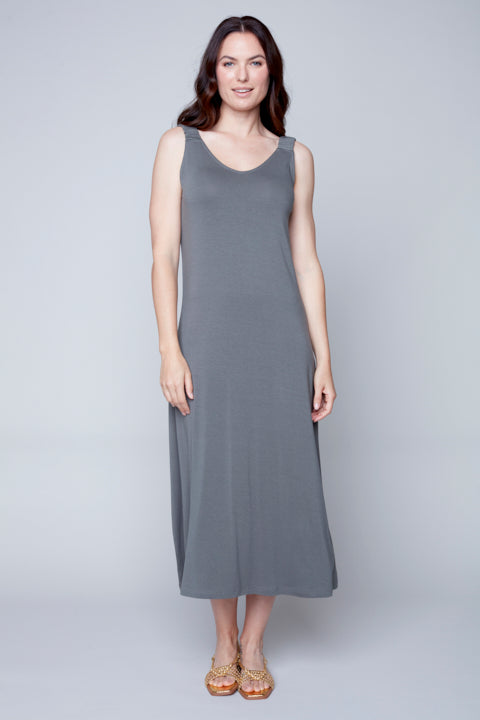 A woman standing against a plain background wearing a Sleeveless Flow Dress by Carre Noir made of soft fabric and embellished sandals.