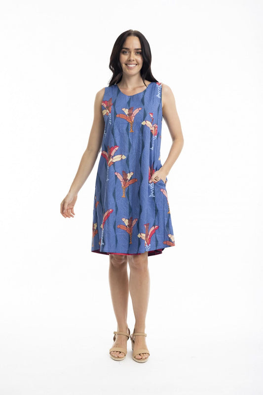 A woman stands in profile wearing the Orientique El Gouna Reversible Shift Dress, a blue dress with red and orange floral patterns and pockets, styled with beige wedge sandals against a plain white background.