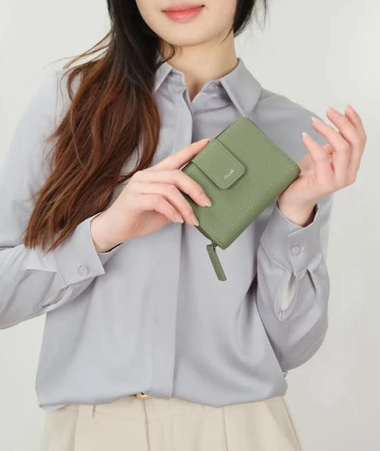 A person in a light gray shirt and beige pants holds The Lucie Small Wallet by Co-Lab, an eco-friendly, compact green wallet with both zipper and snap closures.