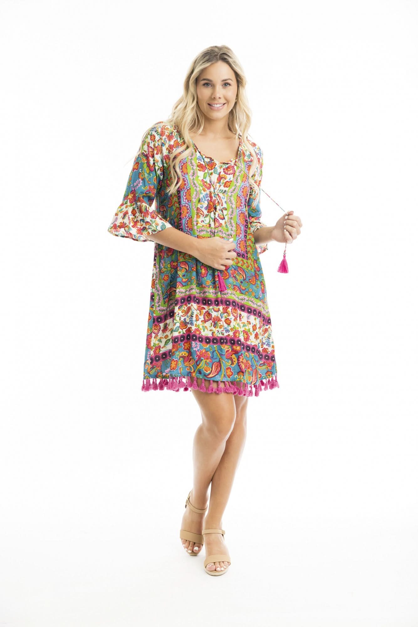 A woman in the vibrant Orientique Carvalho Tassel Dress with bell sleeves and pink tassel ties, paired with beige sandals, stands against a white background—an ideal summer look for warm weather occasions.
