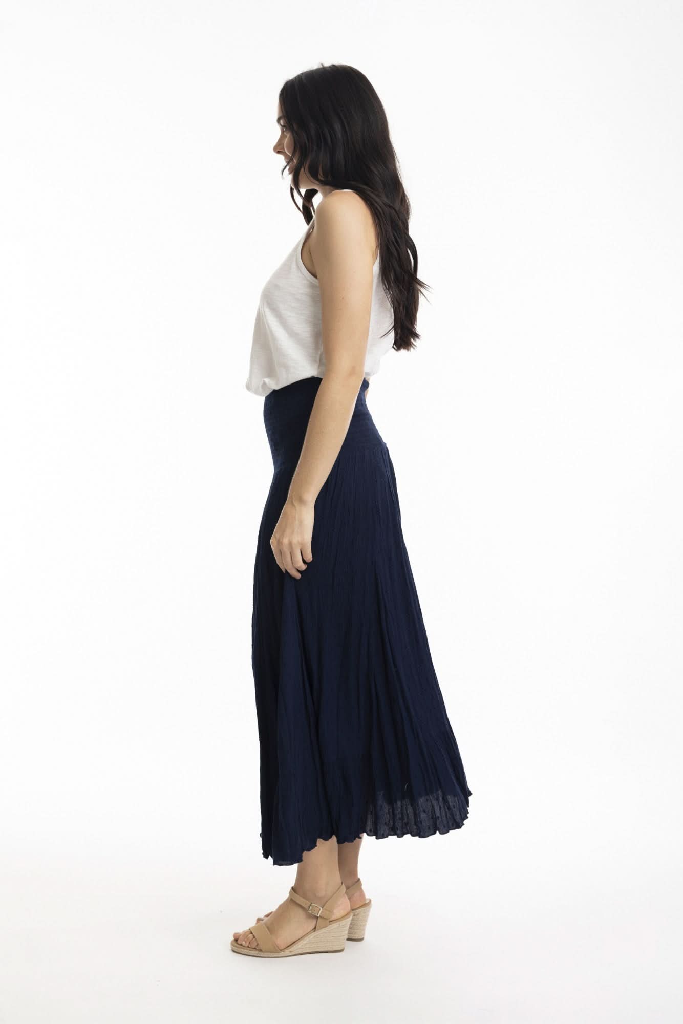 A woman stands against a white background wearing the Orientique Essentials Dobby Skirt Dress— a sleeveless, navy blue, knee-length style — paired with beige sandals and holding the sides of her dress.