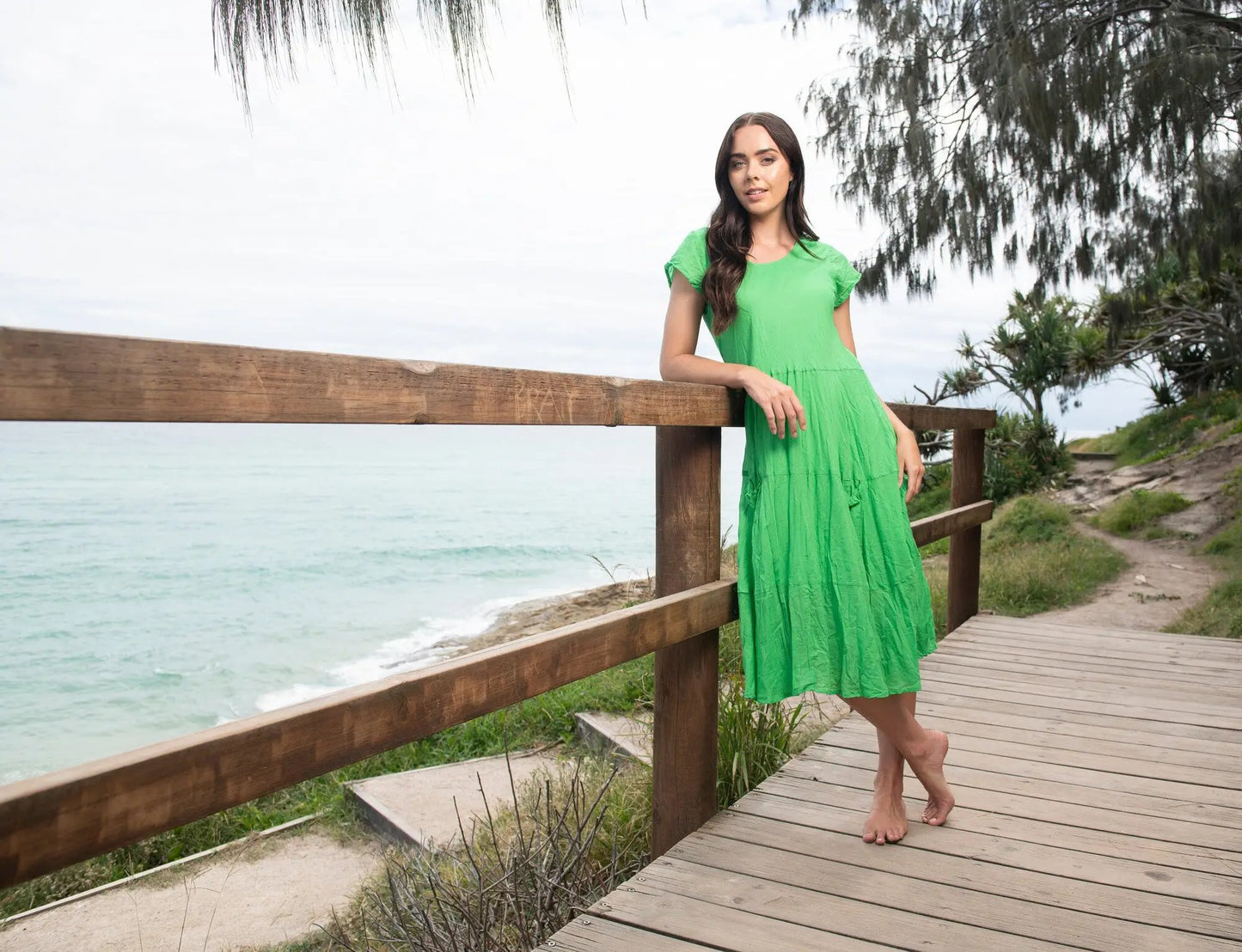A woman in an Orientique Essentials Dress Front Pocket leans on a wooden fence along a beachside boardwalk, with a turquoise sea and overcast sky in the background.