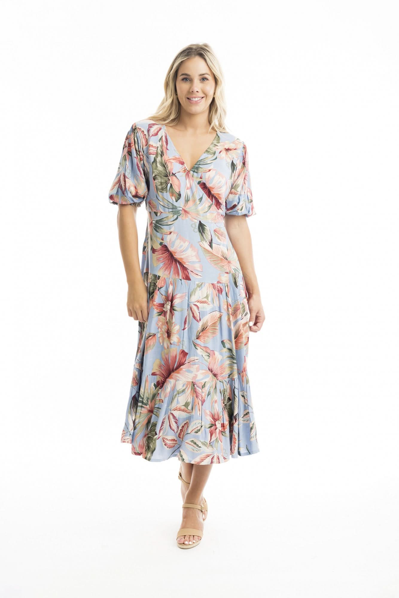 A woman models the Orientique Luzon Midi Crossover Dress—a light blue, floral-patterned summer dress with a v-neckline, short sleeves, and ruffled hem—paired with tan heeled sandals against a white background.