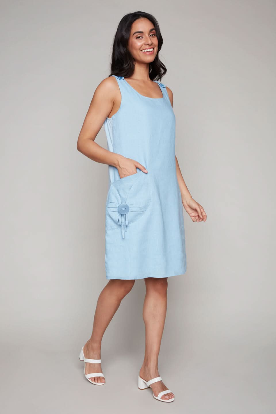 Wearing a Carre Noir Blue Linen Dress with pocket detail, a person smiles as they walk. The high-quality fabric flows gracefully, perfectly paired with white sandals against a neutral backdrop.