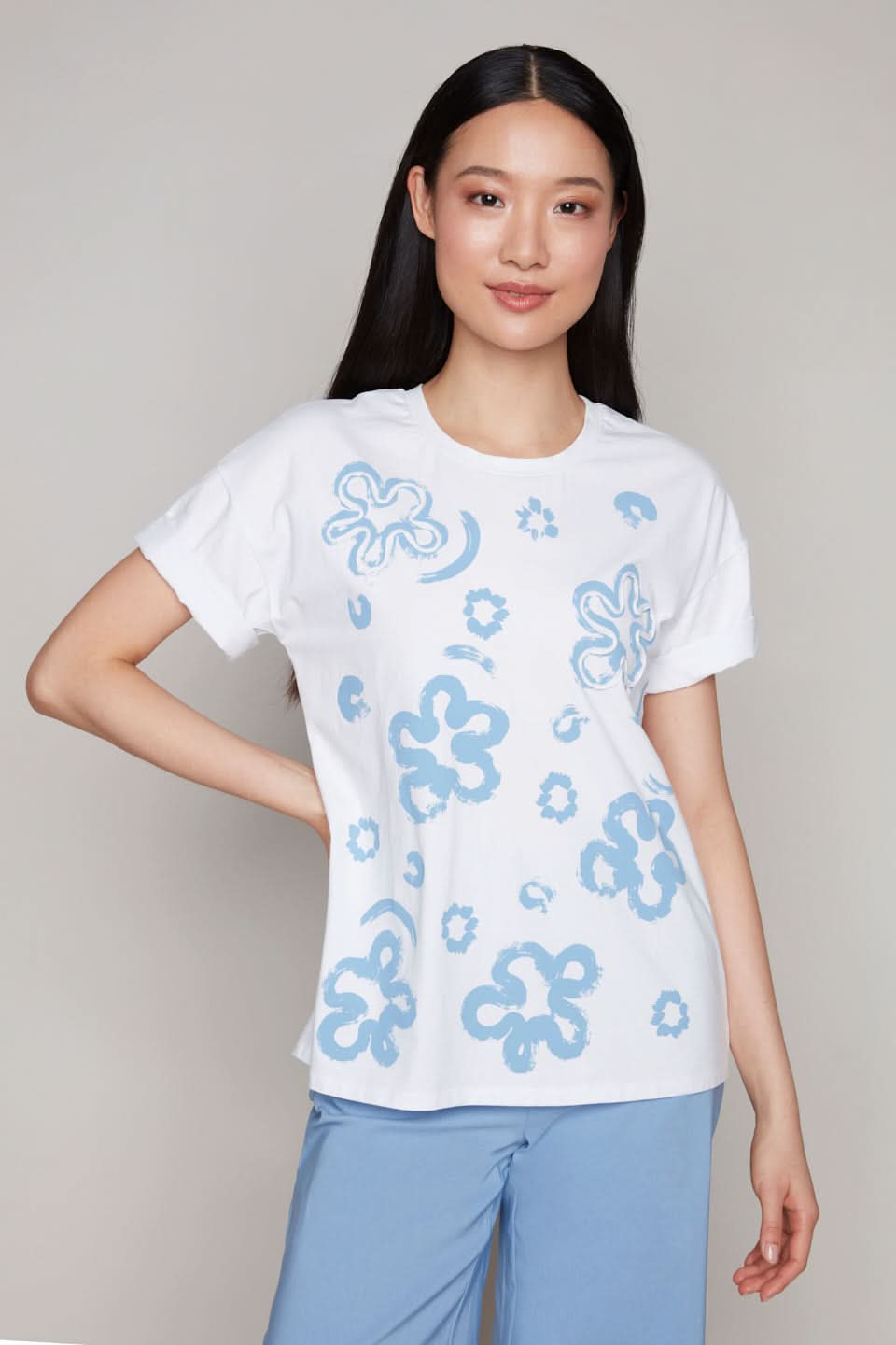 A model presents the Carre Noir Floral Tee with Rolled Cuffs, a timeless wardrobe essential, paired with blue pants against a neutral backdrop.