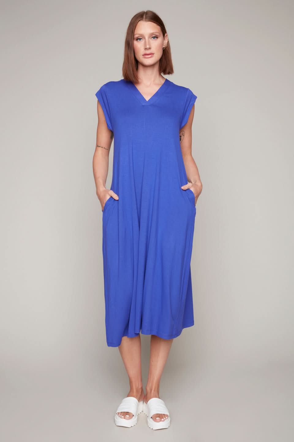 A person stands facing forward wearing the Carre Noir Blue Iris V Neck Dress, made from lightweight fabric with practical pockets, and paired with white sandals against a plain background.