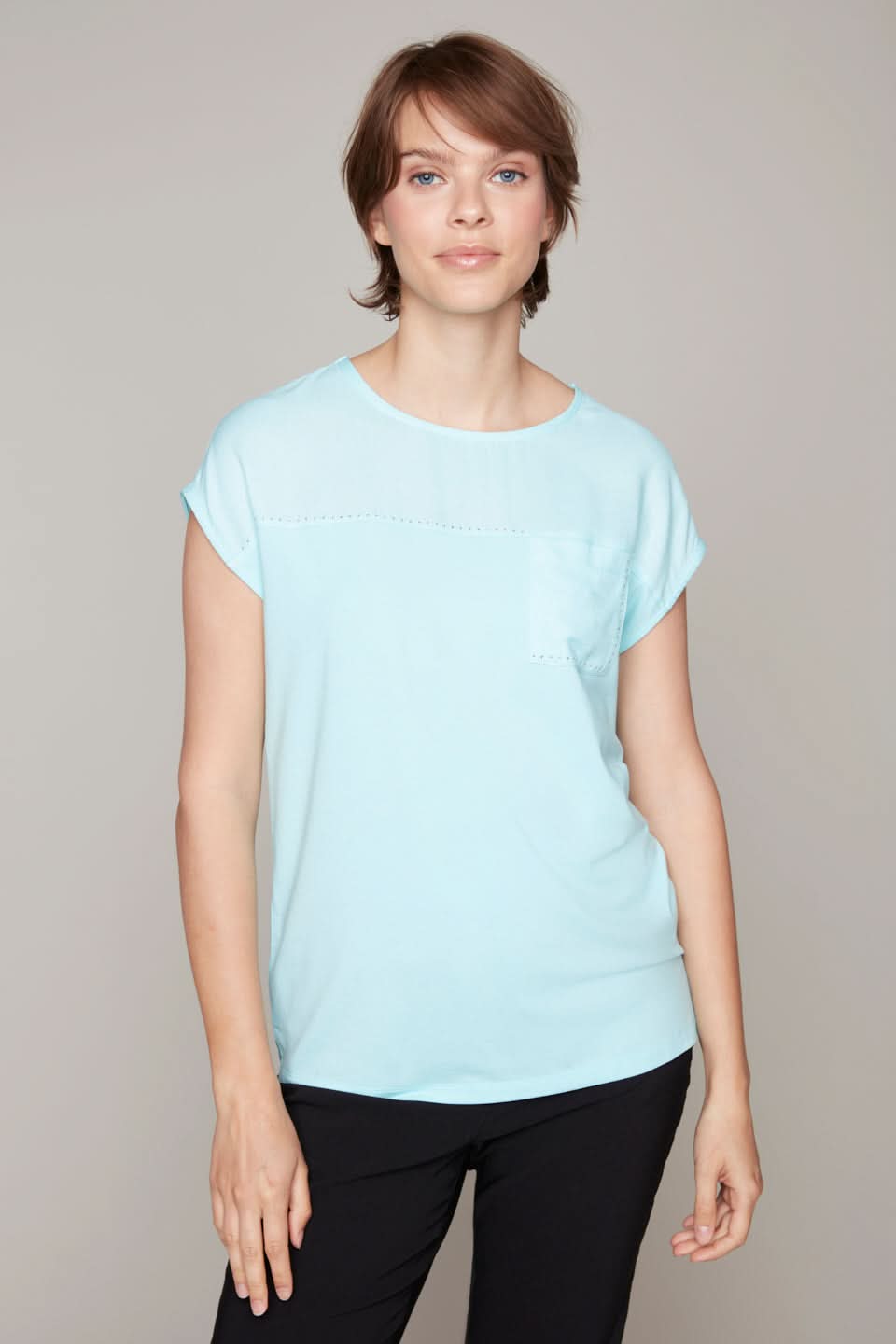 A trendy person with short brown hair, wearing a Carre Noir Aqua Round Neck Top and black pants, stands against a plain background.