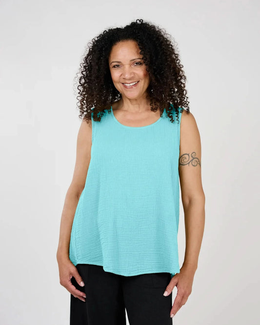 A woman with curly hair smiles brightly, wearing the Katy Tank Top by Shannon Passero and black pants, posing against a light background. Her tattoo on the left arm enhances the colorful vibe of the photo.