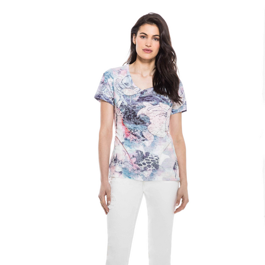 A woman standing against a white background wearing an Orly short sleeve floral tee and white pants.