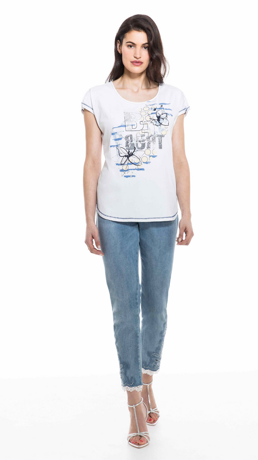 A woman stands facing the camera, wearing a stylish Orly BFF White Tee with a blue graphic design, blue jeans with frayed hems, and silver strappy sandals.