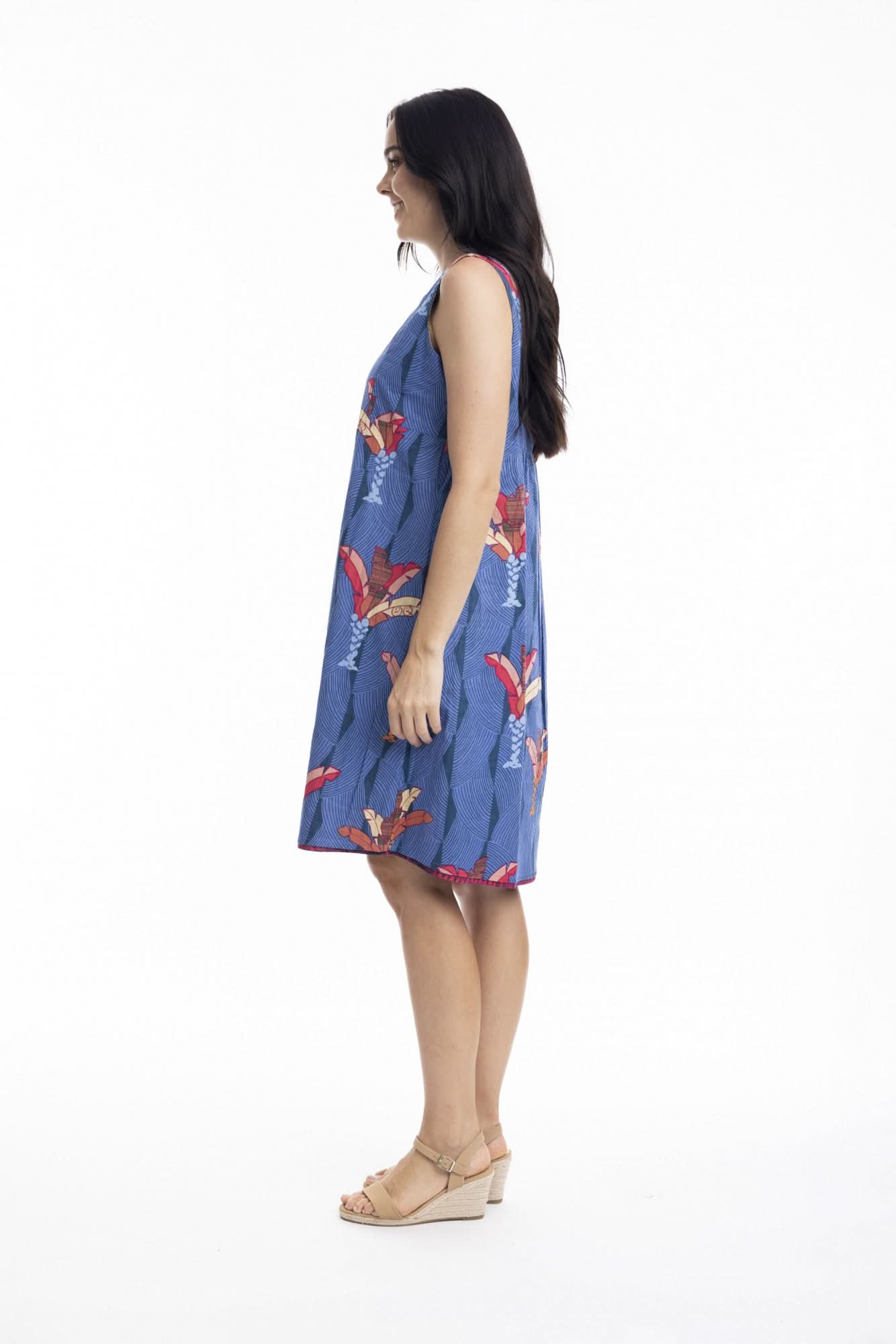 A woman stands in profile wearing the Orientique El Gouna Reversible Shift Dress, a blue dress with red and orange floral patterns and pockets, styled with beige wedge sandals against a plain white background.