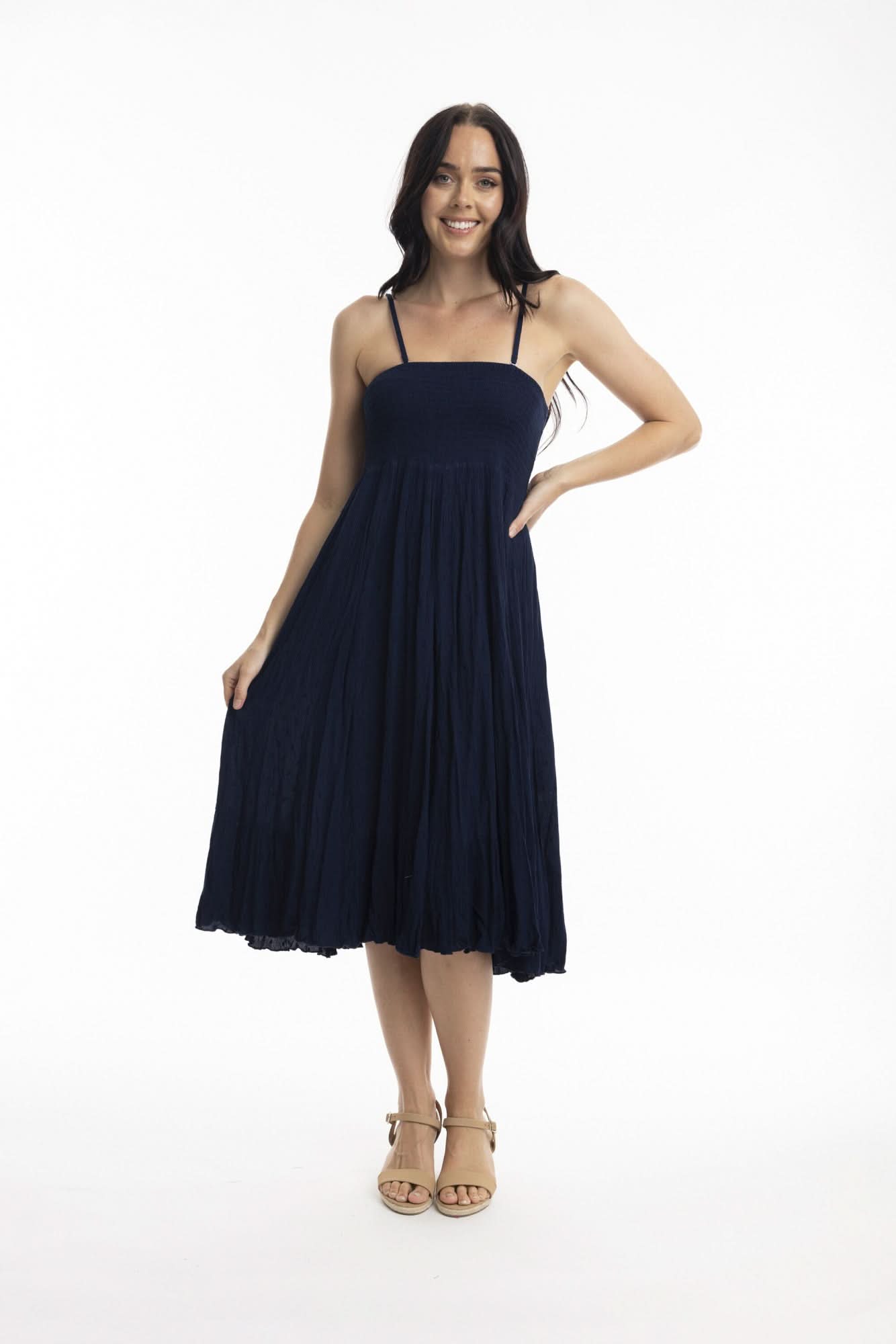 A woman stands against a white background wearing the Orientique Essentials Dobby Skirt Dress— a sleeveless, navy blue, knee-length style — paired with beige sandals and holding the sides of her dress.