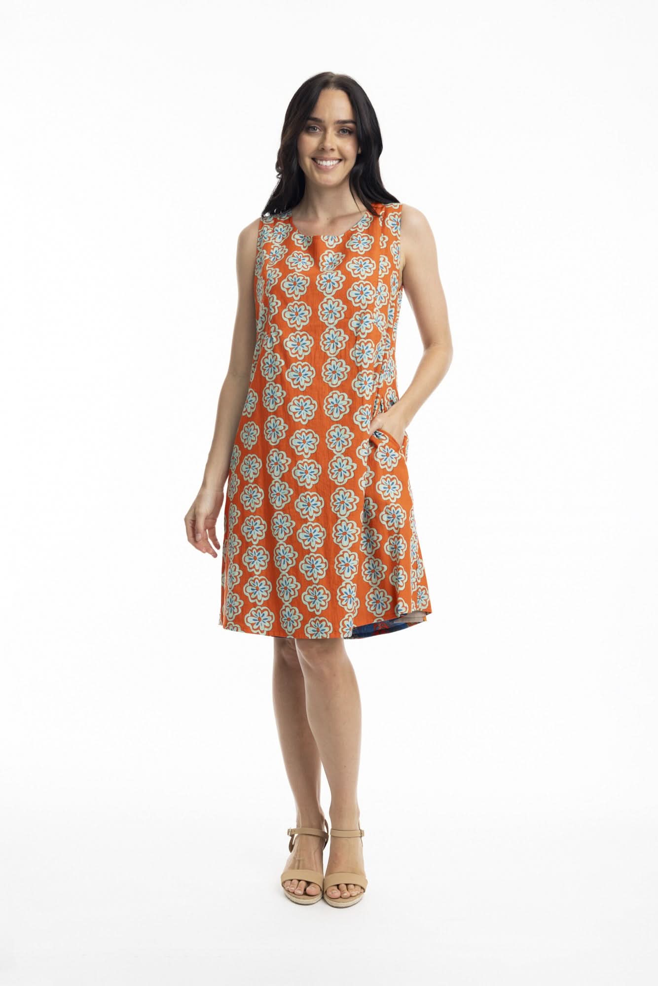 A woman stands smiling in the Orientique Camps Bay Sleeveless Reversible Dress, featuring a vibrant geometric and floral pattern, paired with beige high-heeled sandals against a white background—an ideal summer look.