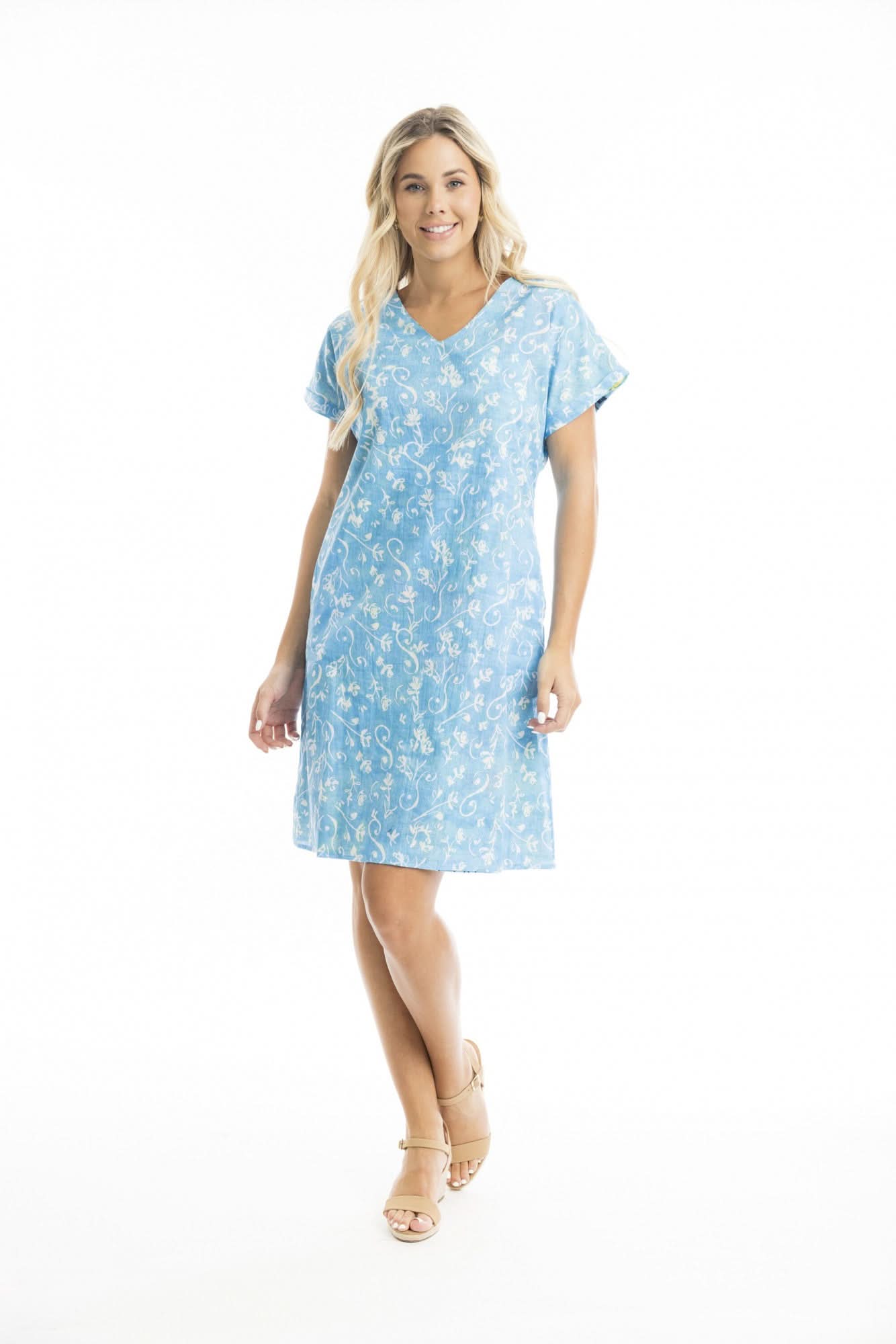 A woman stands against a white background wearing the Orientique Elafonissi Reversible Short Sleeve Dress—a light blue, knee-length V-neck with a white floral pattern and cap sleeves—paired with beige sandals.