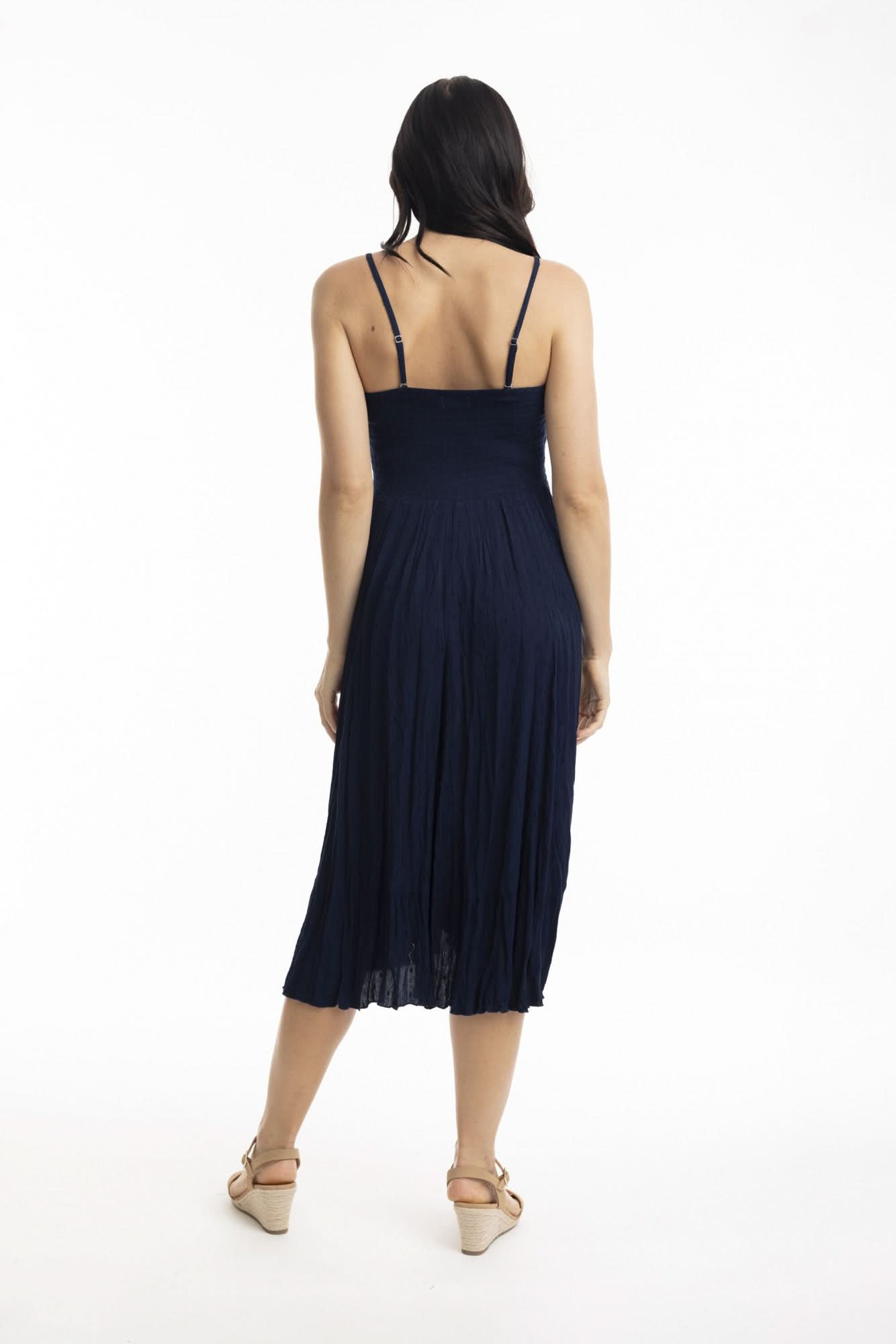 A woman stands against a white background wearing the Orientique Essentials Dobby Skirt Dress— a sleeveless, navy blue, knee-length style — paired with beige sandals and holding the sides of her dress.