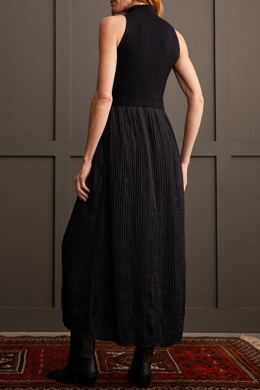 A person wearing Tribal's Sleeveless Twofer Maxi Dress, which features a high neck and a long, pleated skirt, stands against a gray paneled wall. They are also wearing black boots.